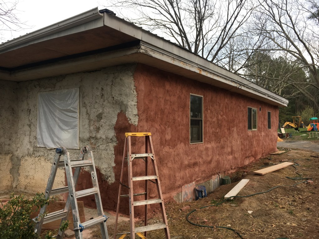 Side of house during stuccoing -- some with grey first coat, some with red second coat of stucco, and ladders in front.